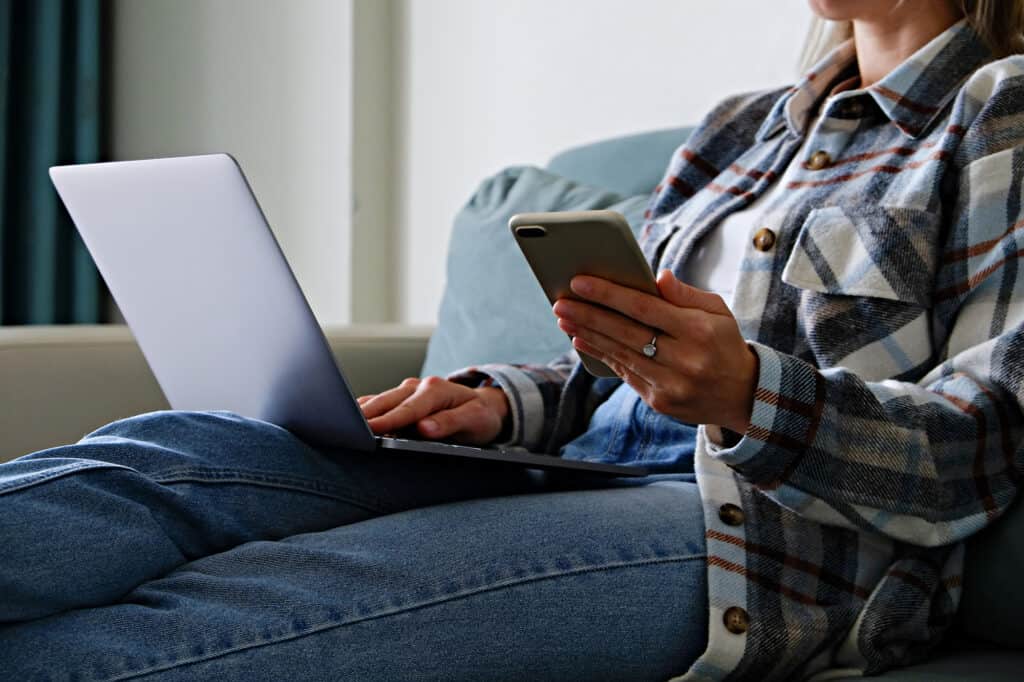 A woman sitting on a couch with a laptop and cell phone, while starting a business podcast.