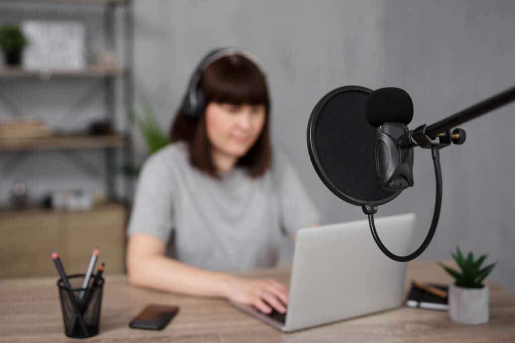 A woman is sitting at a desk with a microphone and laptop, hosting her business podcast.