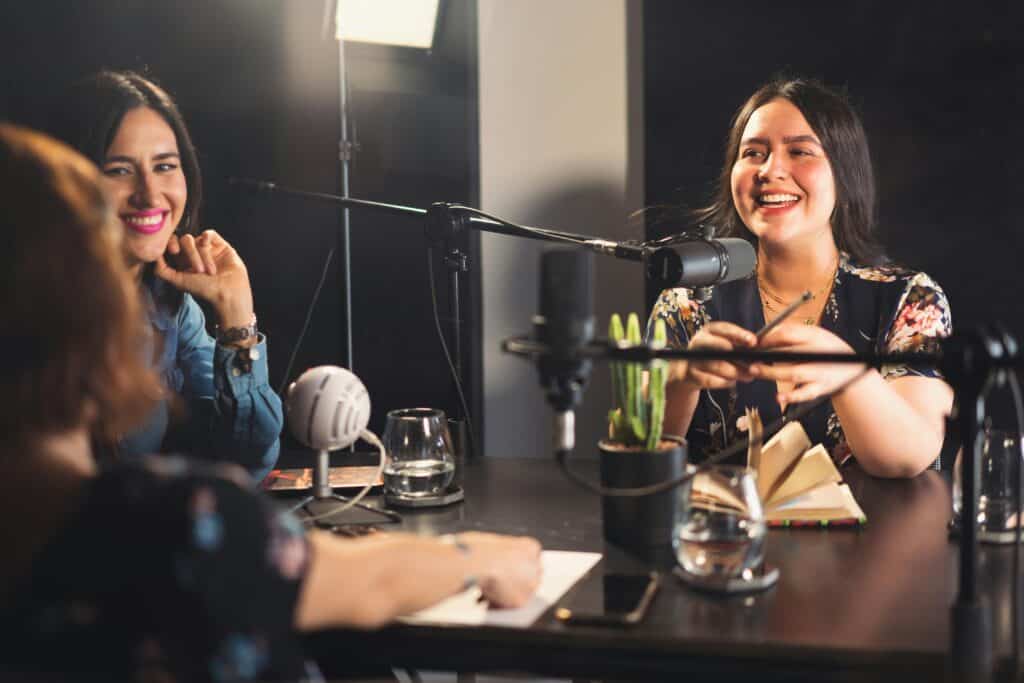 Three women sitting at a table with microphones in front of them, recording for their podcast startup.