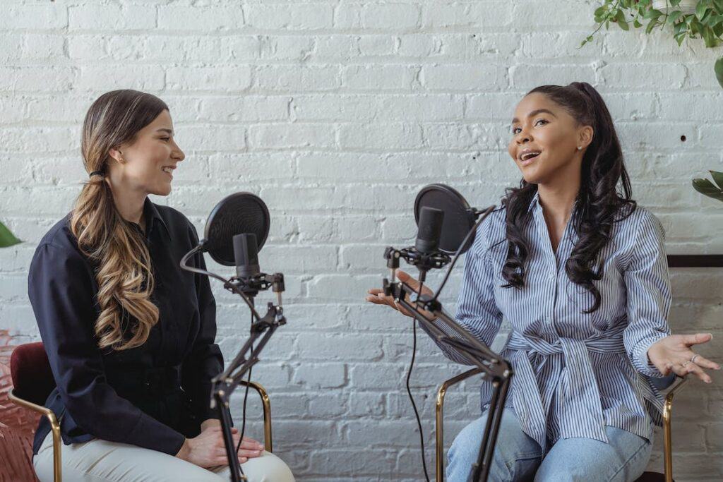 Two women podcasting on a microphone in front of a brick wall while discussing their business plan.
