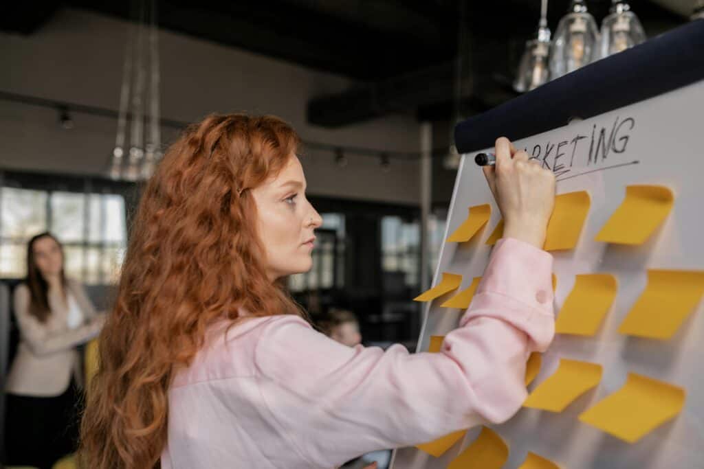 A woman writing on a whiteboard with sticky notes for a digital product selling course.