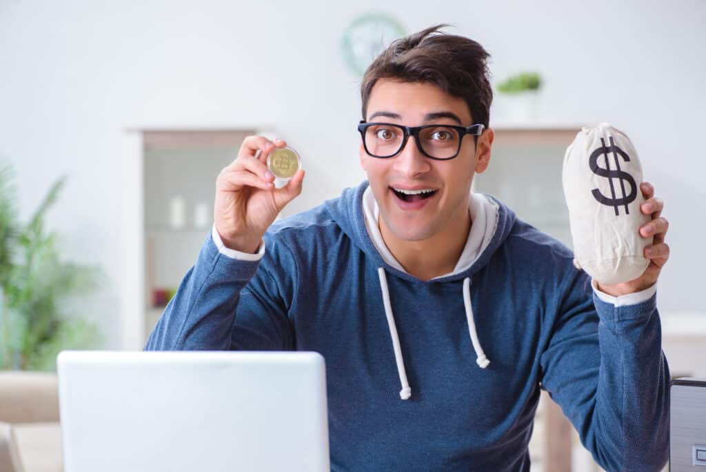Man holding a coin and a bag of money appears excited in front of his laptop exploring active income vs passive income.