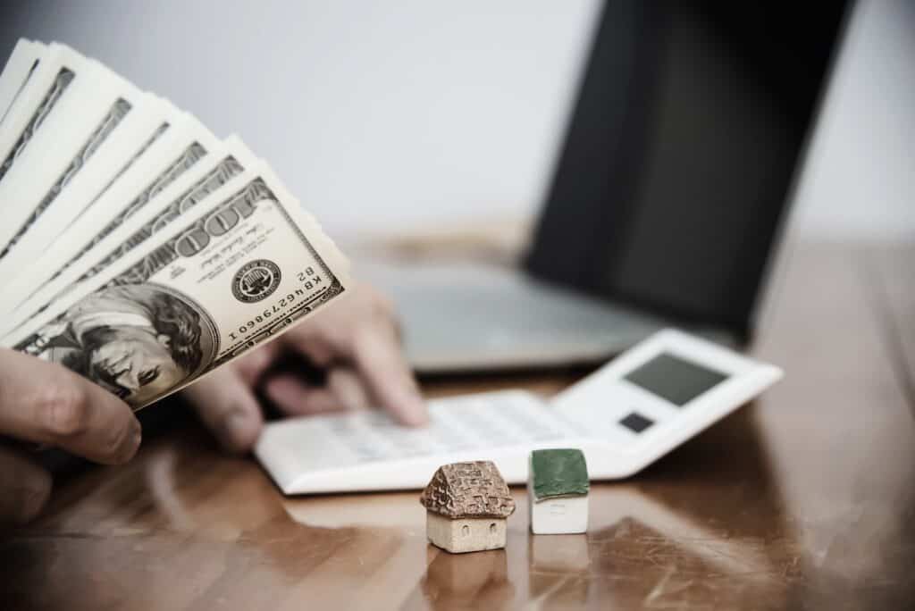 A person using a calculator near a laptop, with a bundle of US dollar bills in hand and miniature houses on the table, possibly calculating passive income from real estate investments or mortgage.