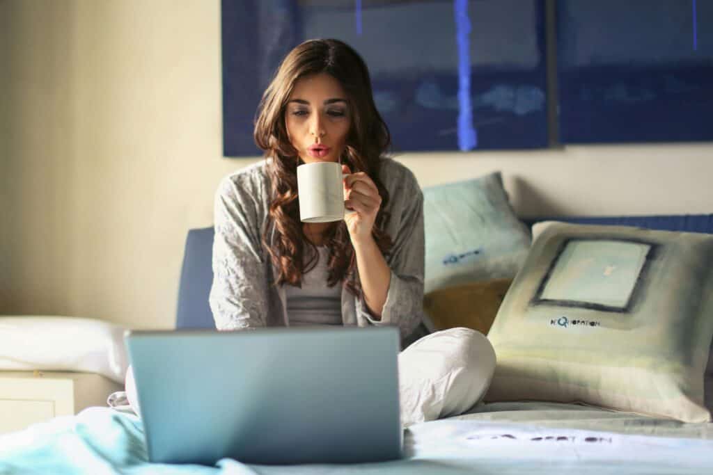 Woman sitting on a bed, drinking coffee, and looking at a laptop screen to learn how to make residual income from home.
