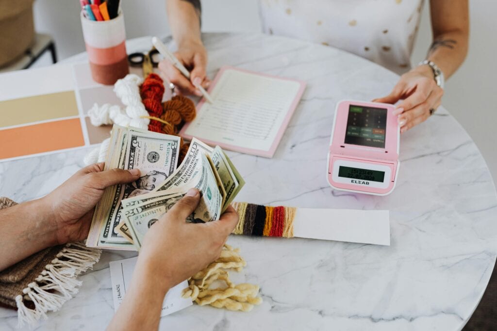 Two individuals are engaged in a craft-related transaction, focusing on how to make residual income from home; one person counts cash while the other records details, with yarn and a clock visible on a marble table