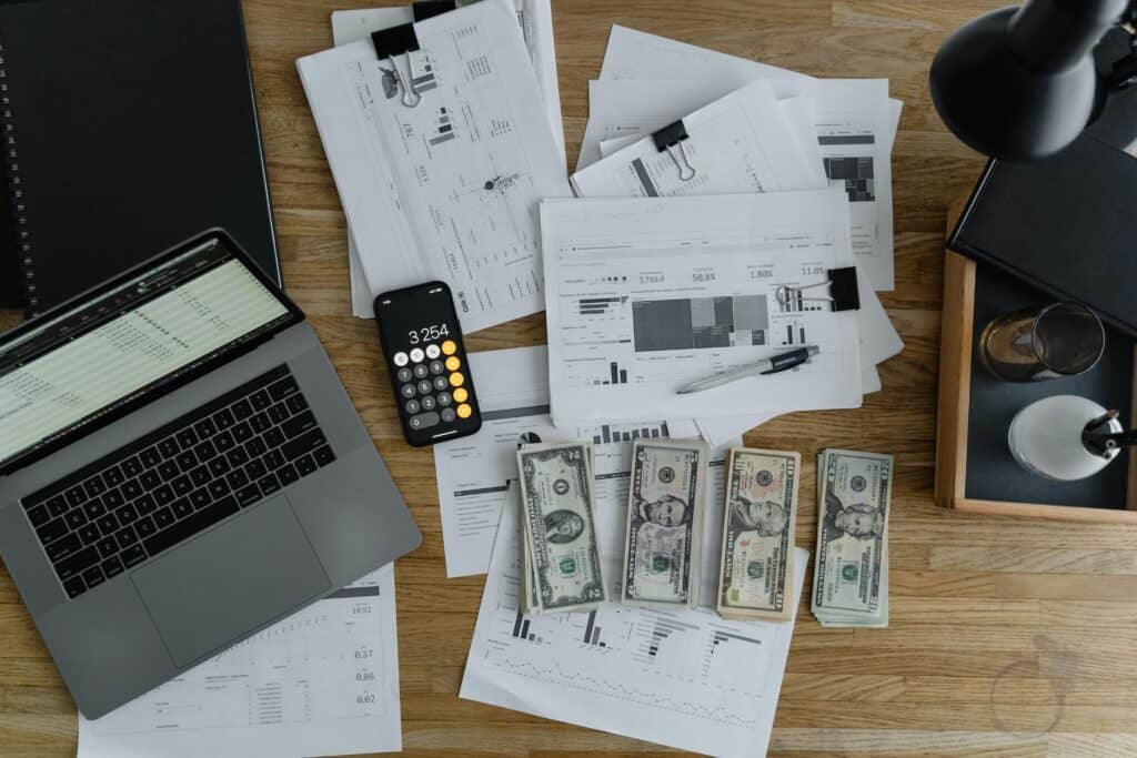 A laptop, financial documents related to passive income businesses to buy, a calculator, and cash spread out on a wooden desk.