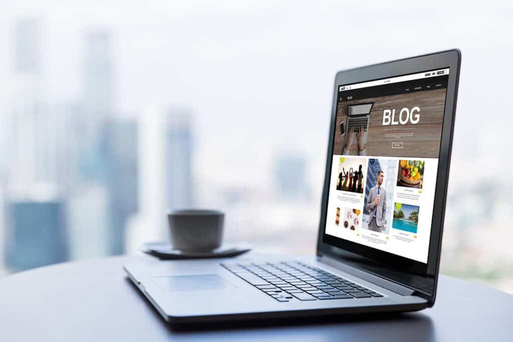 A laptop displaying a blogging for business webpage is placed on a table with a cup and saucer in the background. The image is taken in front of a window with an out-of-focus cityscape view.