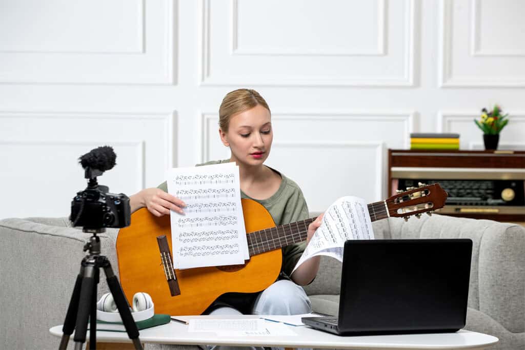 A person sits on a sofa holding a guitar and sheet music, with a laptop and a camera in front. They appear to be recording or streaming their session, possibly blogging for business purposes.