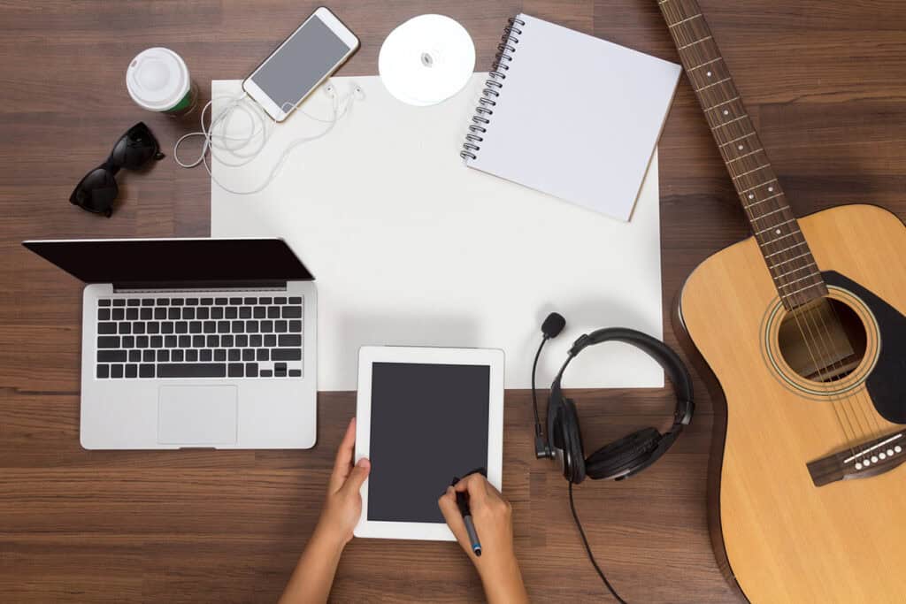 A workspace with a laptop, tablet, smartphone, earphones, notebook, pen, CD, coffee cup, sunglasses, and an acoustic guitar on a wooden table. A person uses a stylus on the tablet while brainstorming ideas for blogging for business.
