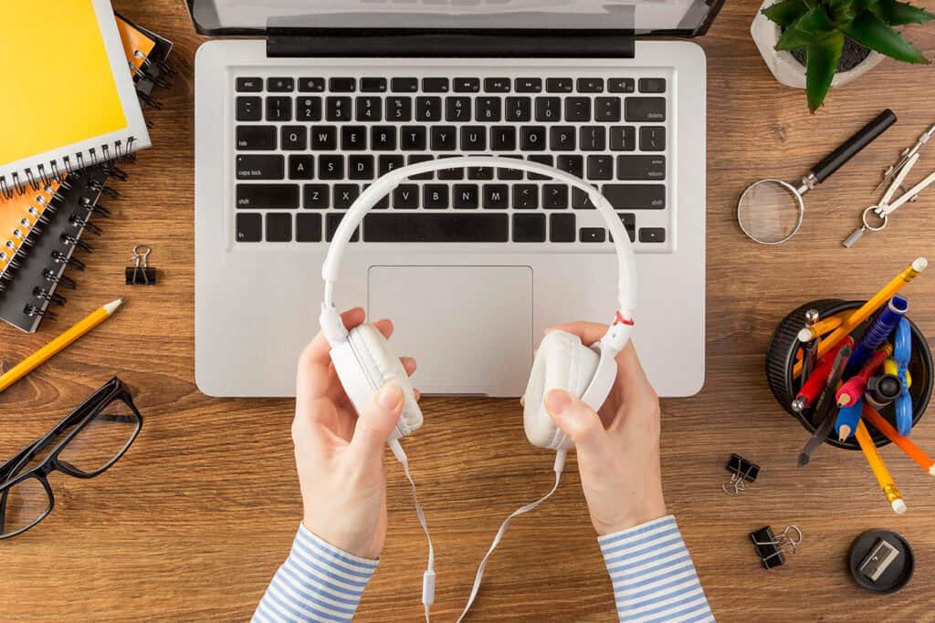A person holds white headphones above a laptop on a wooden desk, surrounded by notebooks, glasses, clips, pens, and a plant—an ideal setup for blogging for business.