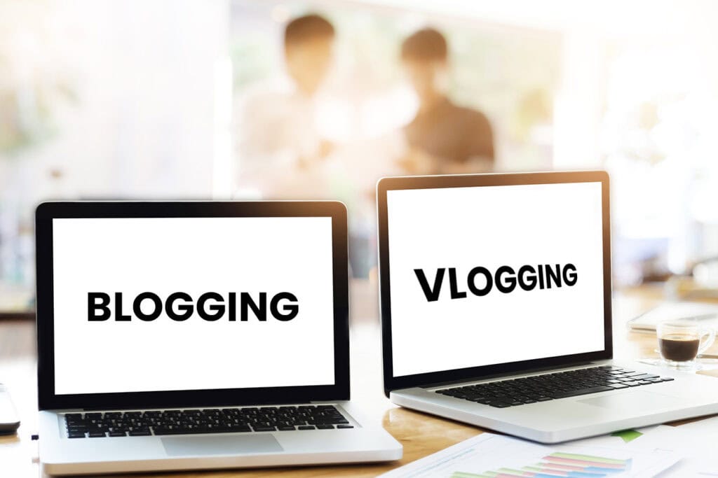 Two laptops on a desk display the words "BLOGGING" and "VLOGGING" on their screens, with two blurred people in the background, highlighting the importance of blogging for business.