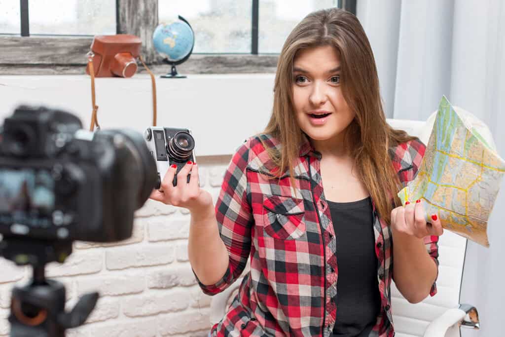 A person is seated indoors holding a camera and a map, with another camera positioned in front of them. They appear to be speaking or explaining something, possibly sharing tips on blogging for business.