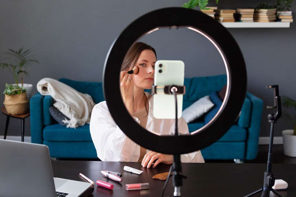 A person applies makeup in front of a smartphone mounted on a ring light, sitting at a desk with various makeup products and a laptop, seamlessly blending their passion with blogging for business.