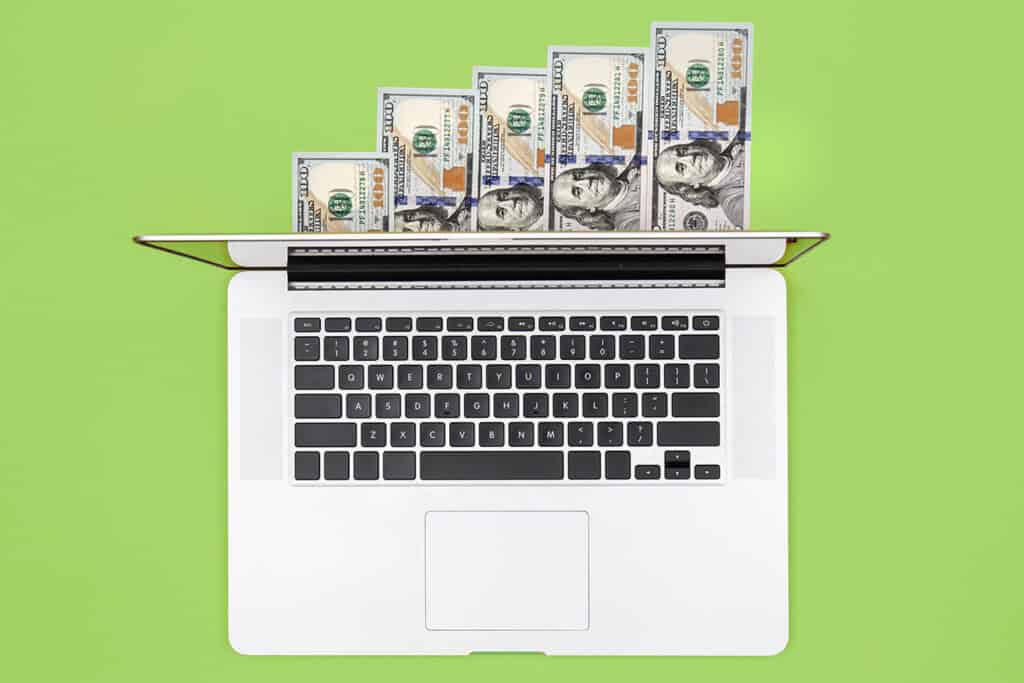 A laptop with several U.S. $100 bills partially inserted into the screen, set against a green background, symbolizing the potential earnings from blogging for business.
