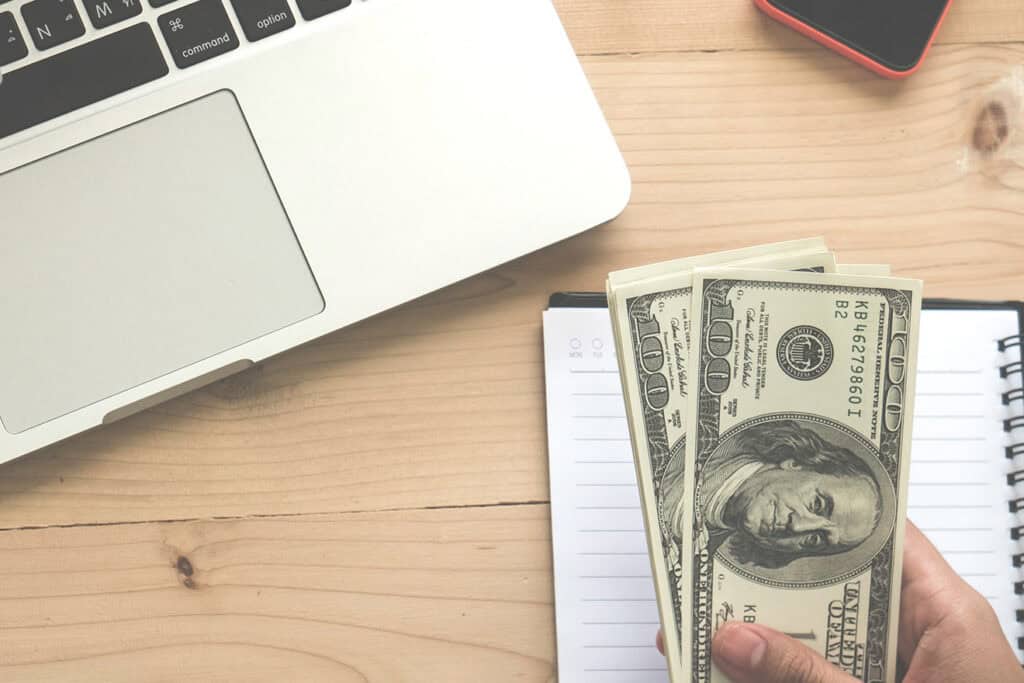 A hand holding a stack of US hundred-dollar bills next to a silver laptop, a red smartphone, and an open notebook on a wooden desk—an ideal setup for blogging for business.