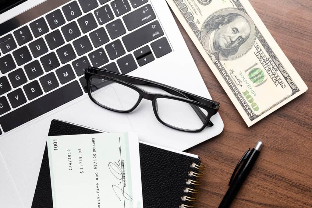 A pair of glasses, a check, and a pen rest on a laptop keyboard next to a $100 bill and a notebook on a wooden surface—perfect tools for blogging for business.
