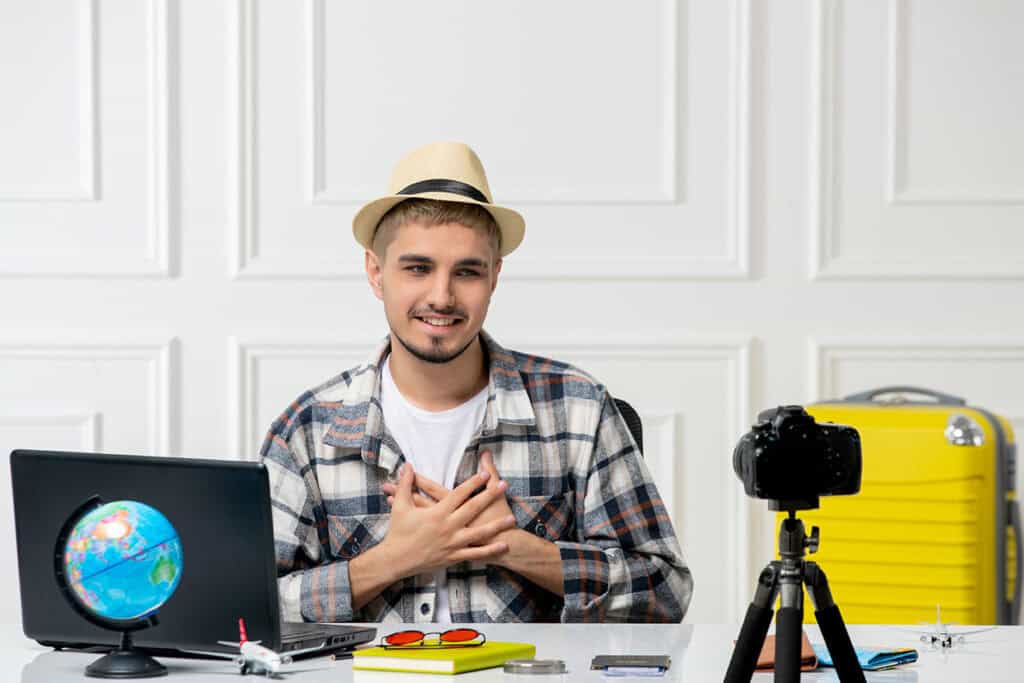 A man wearing a hat and plaid shirt sits at a desk with a laptop, camera, model airplanes, and a yellow suitcase, speaking and gesturing towards the camera about blogging for business.