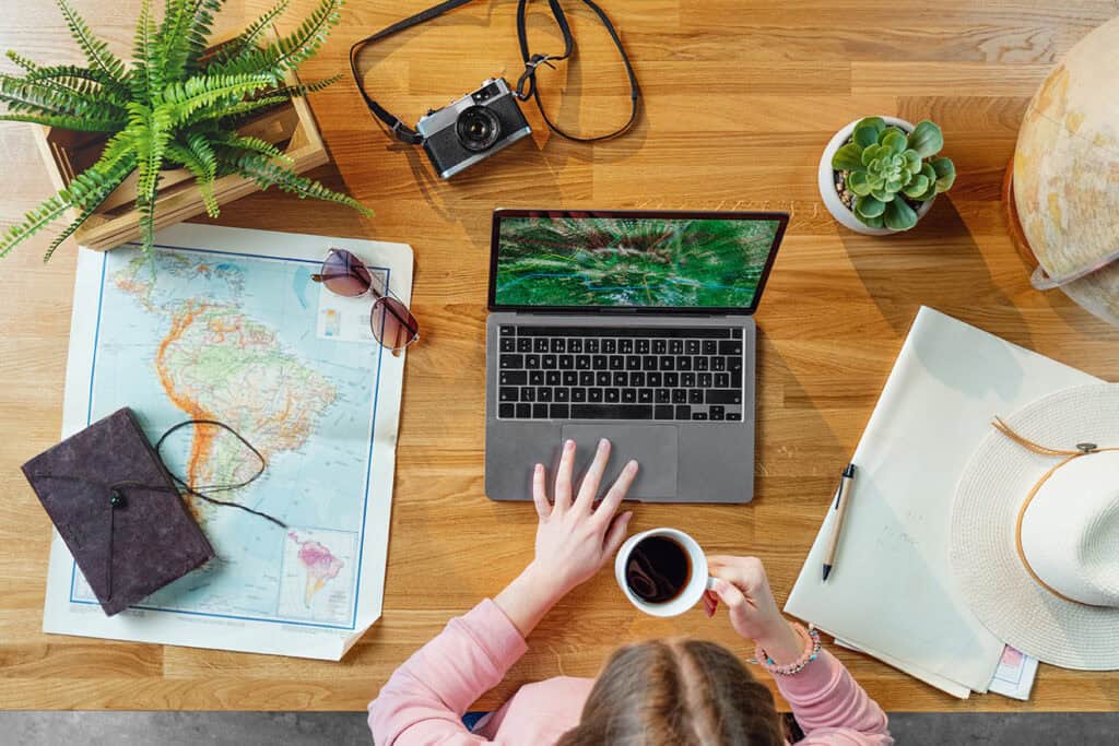 A person holds a coffee cup while using a laptop at a wooden desk containing a world map, sunglasses, a hat, a camera, plants, and a notebook—an ideal setup for blogging for business.
