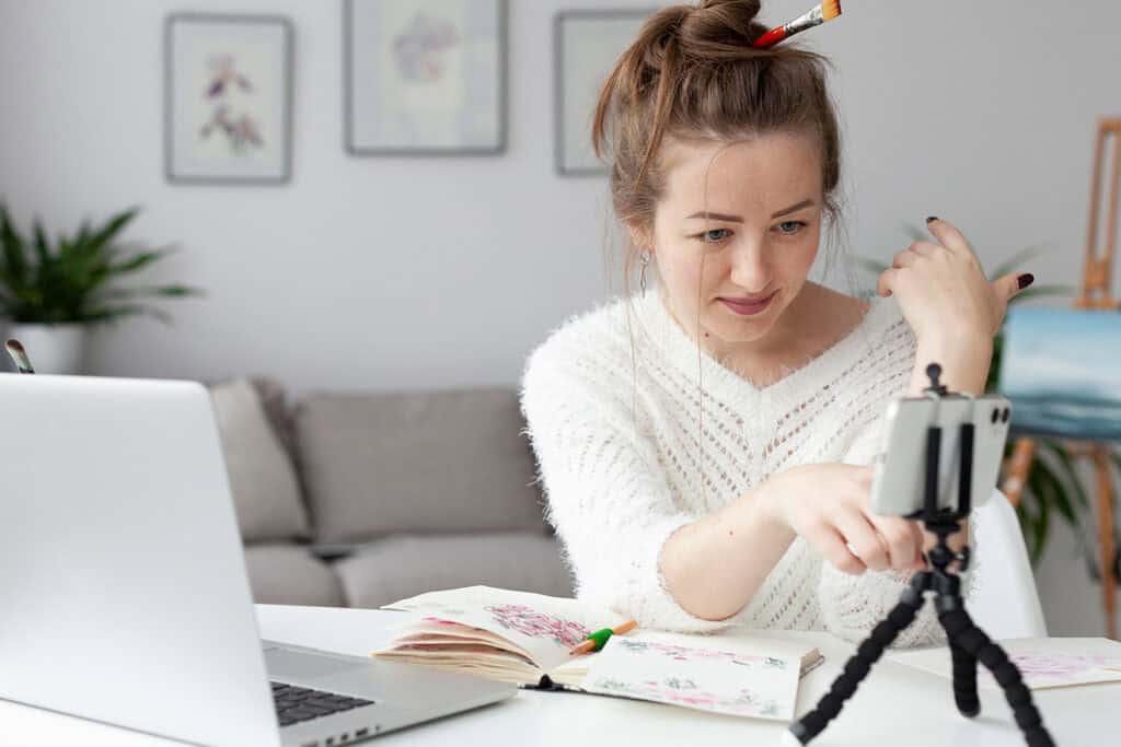A woman with a pencil in her hair interacts with her smartphone on a tripod while working at a desk with an open notebook, laptop, and art supplies in a home setting, perhaps focusing on blogging for business.