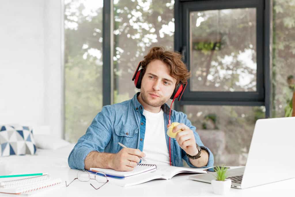 A man wearing headphones sits at a desk with an open notebook and laptop, holding a pen and an apple, appearing deep in thought, perhaps contemplating his next post on blogging for business.